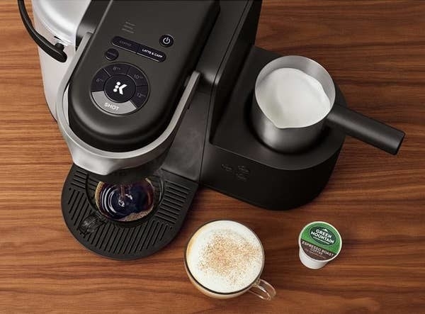 The coffee maker brewing a cup of coffee while frothing milk with a cup of cappuccino and a K-cup sitting next to it