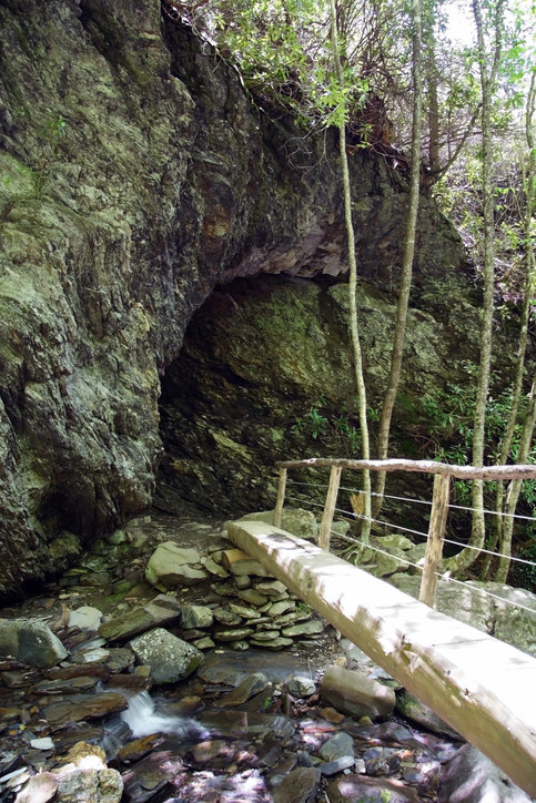 A thin, wooden bridge leads over a stream to a rocky cave entrance 