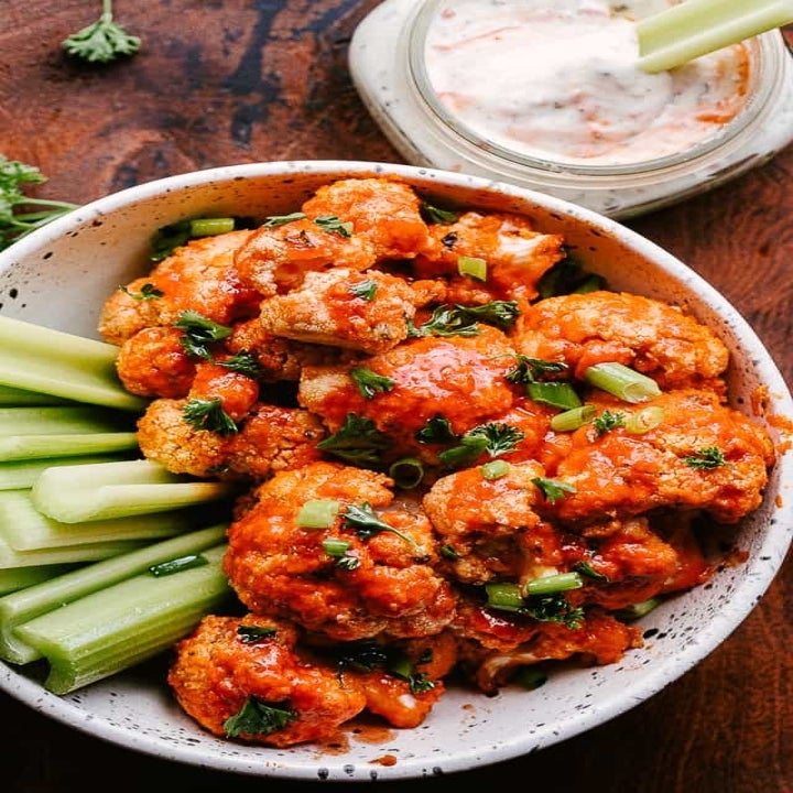 Buffalo cauliflower on a plate with celery sticks, scallions, and blue cheese dipping sauce on the side.