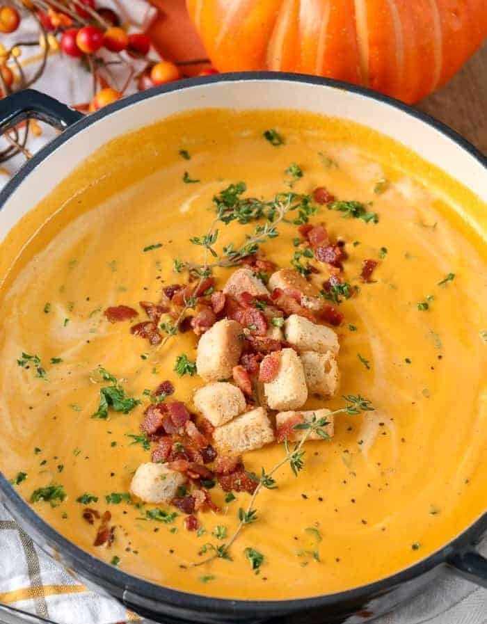 A bowl of creamy pumpkin soup topped with bacon, croutons, and herbs.