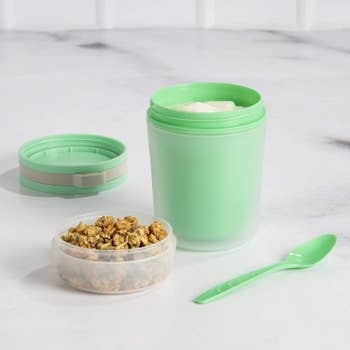 The green container with the clear plastic top removed and being used as a bowl for granola, with yogurt int he base and the spoon and middle piece off to the side
