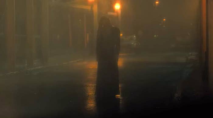 The silhouette of a woman in a long dress and long hair is visible in a rainy alleyway
