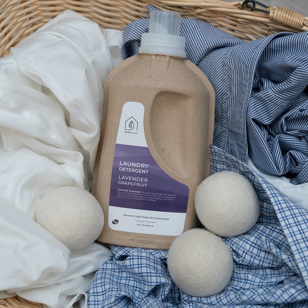 Cardboard container of PUR Home lavender grapefruit laundry detergent with white dryer balls in a laundry basket of clothes