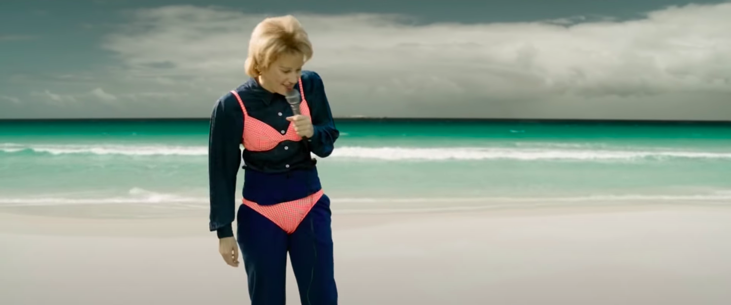Kate McKinnon, as Ellen DeGeneres, does stand up on the beach in a full outfit with a bikini on top of it