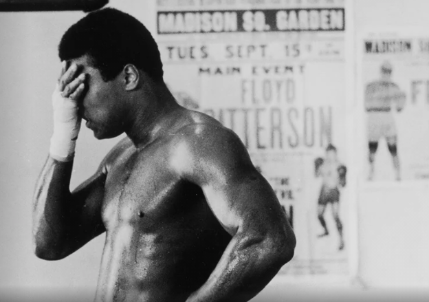 black and white photo of Muhammad Ali shirtless with his right hand on his forehead 