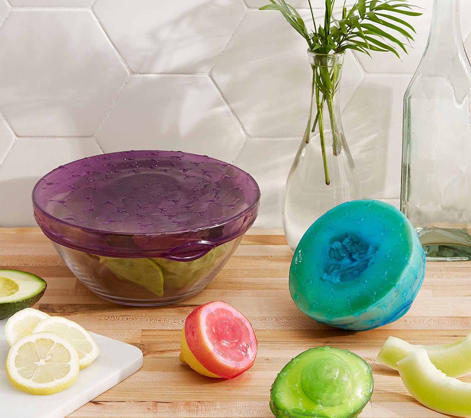 A large glass bowl, along with a half cut lemon, melon, and avocado are covered in a stretchy silicone cover