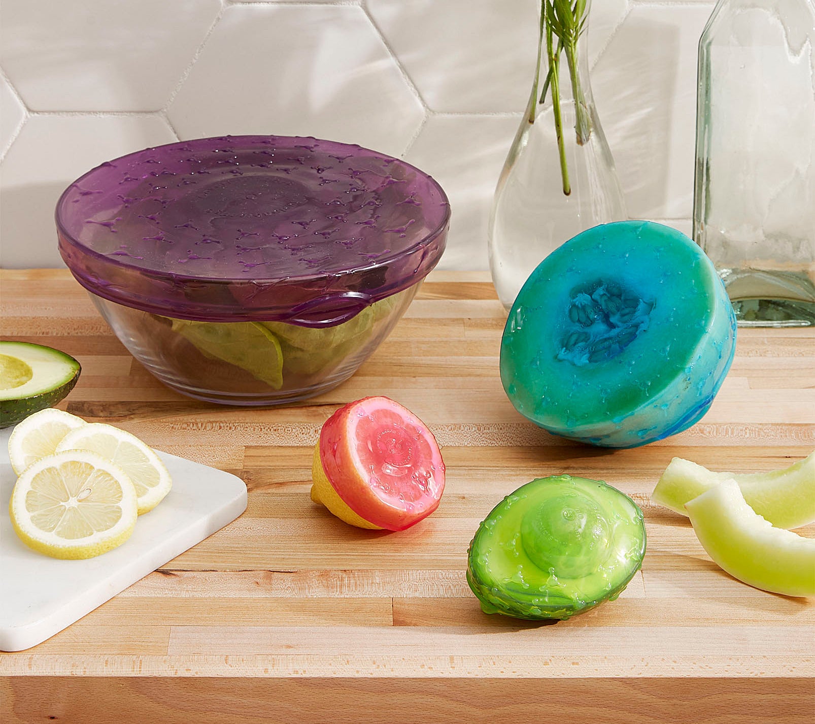 A large glass bowl, along with a half cut lemon, melon, and avocado are covered in a stretchy silicone cover