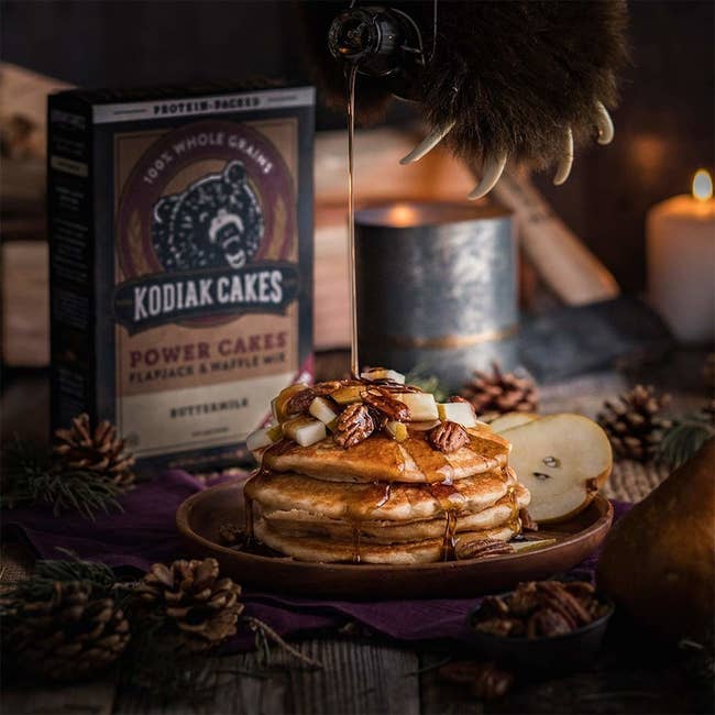 A bear paw pours syrup over a stack of the pancakes, with the Kodiak Power Cakes in the background