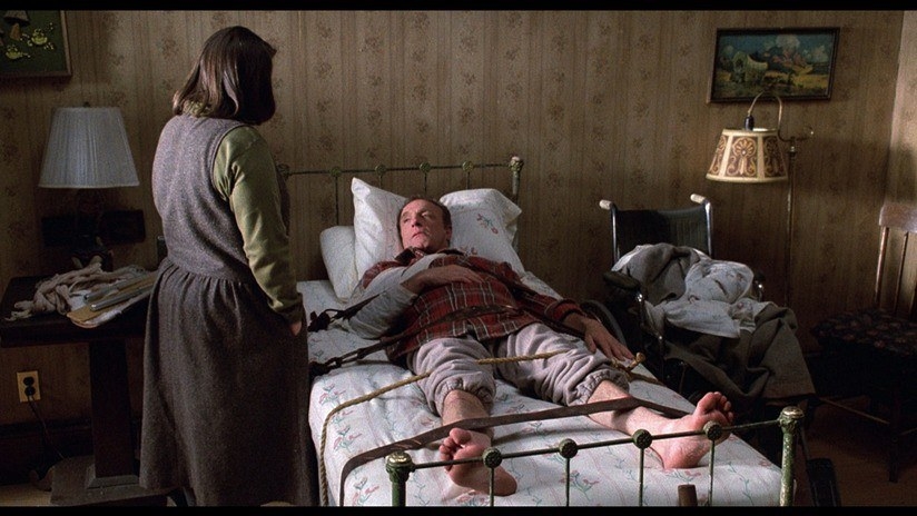 Author, Paul Sheldon, in bed as Annie Wilkes stands over him with a creepy aura
