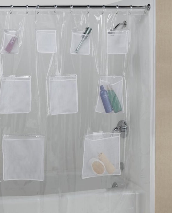 The clear shower curtain featuring varying sizes of pockets holding things like shampoo bottles and a brush, covering a shower/bath