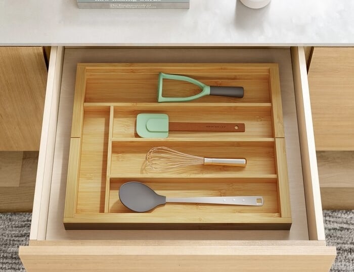 The wooden organizer inside of a drawer holding utensils like a spatula, a whisk, and a spoon