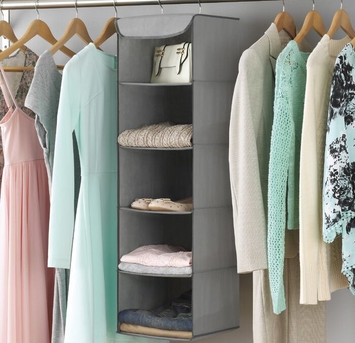 The gray tiered organizer hanging in a closet holding folded pants, folded sweaters, a pair of shoes, and a handbag