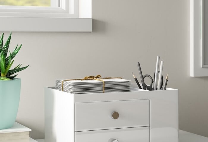 The organizer in white — there are two drawers, a storage cup area filled with pens, pencils, and a scissor, and an additional storage space at the top where a stack of papers are sitting