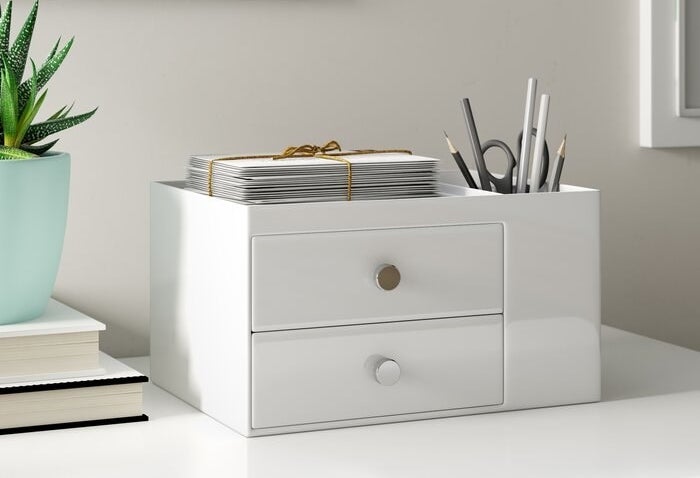 The organizer in white — there are two drawers, a storage cup area filled with pens, pencils, and a scissor, and an additional storage space at the top where a stack of papers are sitting