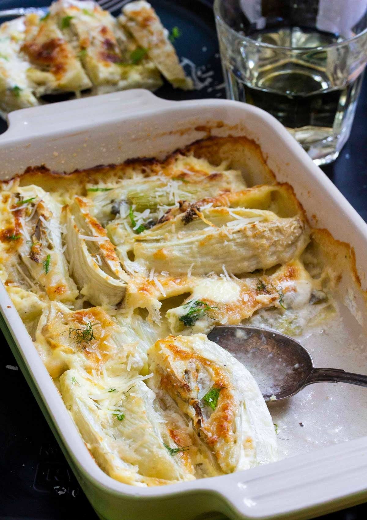 A spoon taking a scoop from a baking pan filled with creamy baked fennel.
