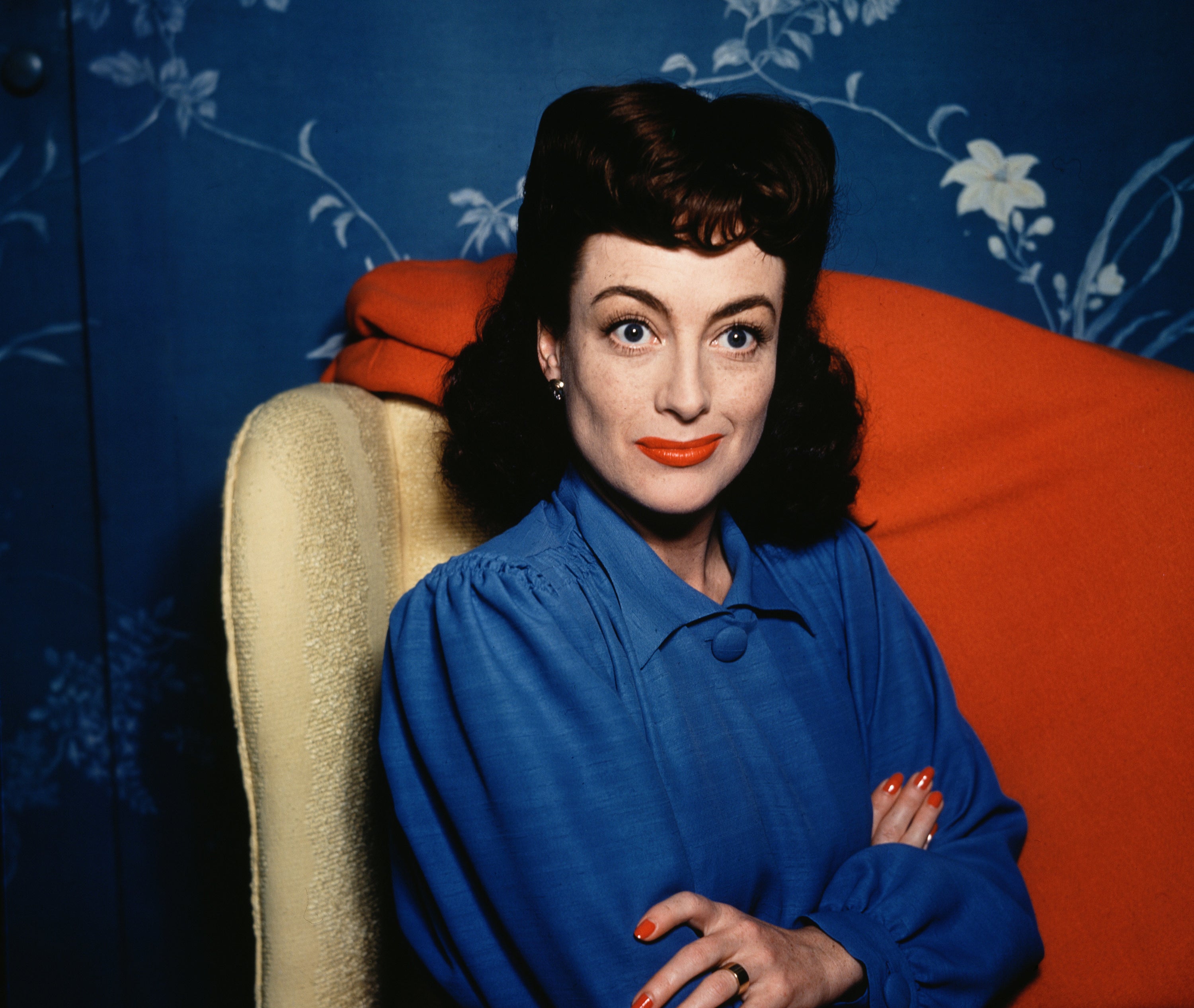 A color photo of Joan Crawford from the 1940s of her sitting an gold armchair with her arms crossed, eyebrows arched, and slightly smiling, while wearing a blue blouse