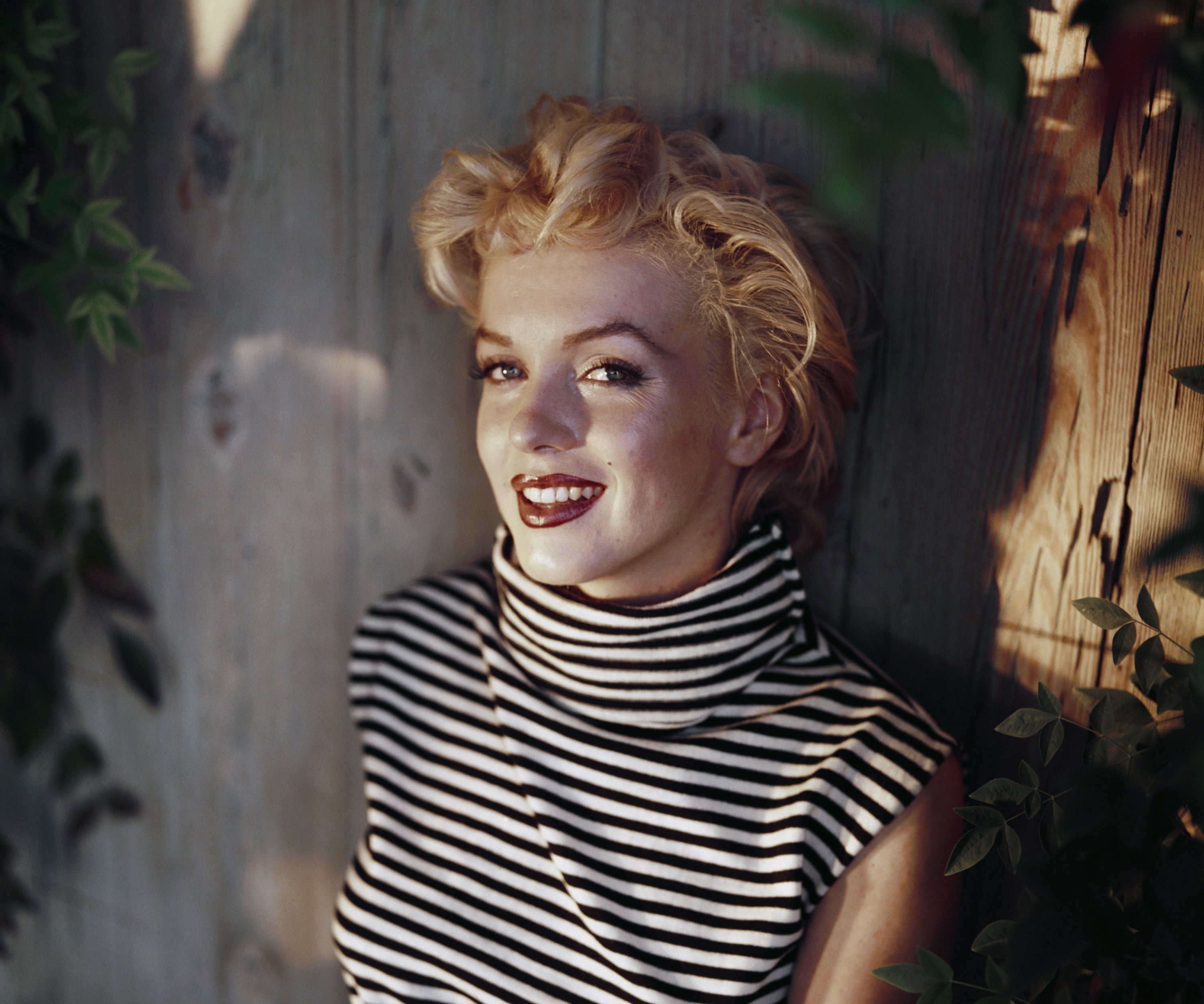 A color photo of Marilyn Monroe in the early &#x27;50s smiling and sitting in a back yard wearing a white and black sleeveless turtleneck