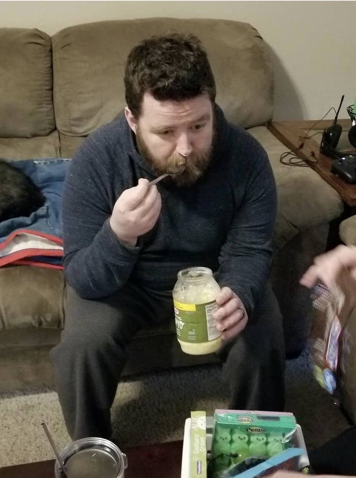 A bearded man sits on a couch eating what looks like mayo from a jar