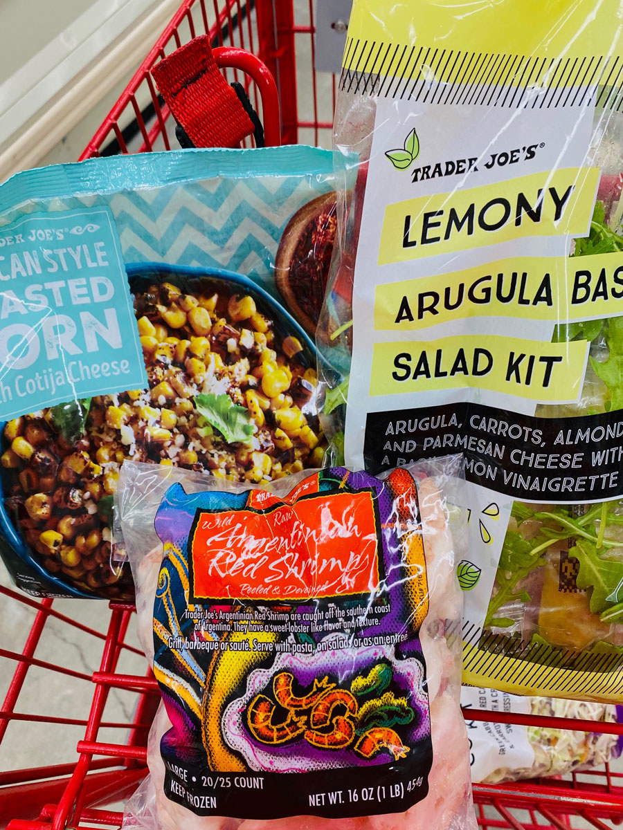 A bag of frozen roasted corn, frozen shrimp, and a lemony arugula salad kit in a shopping cart.