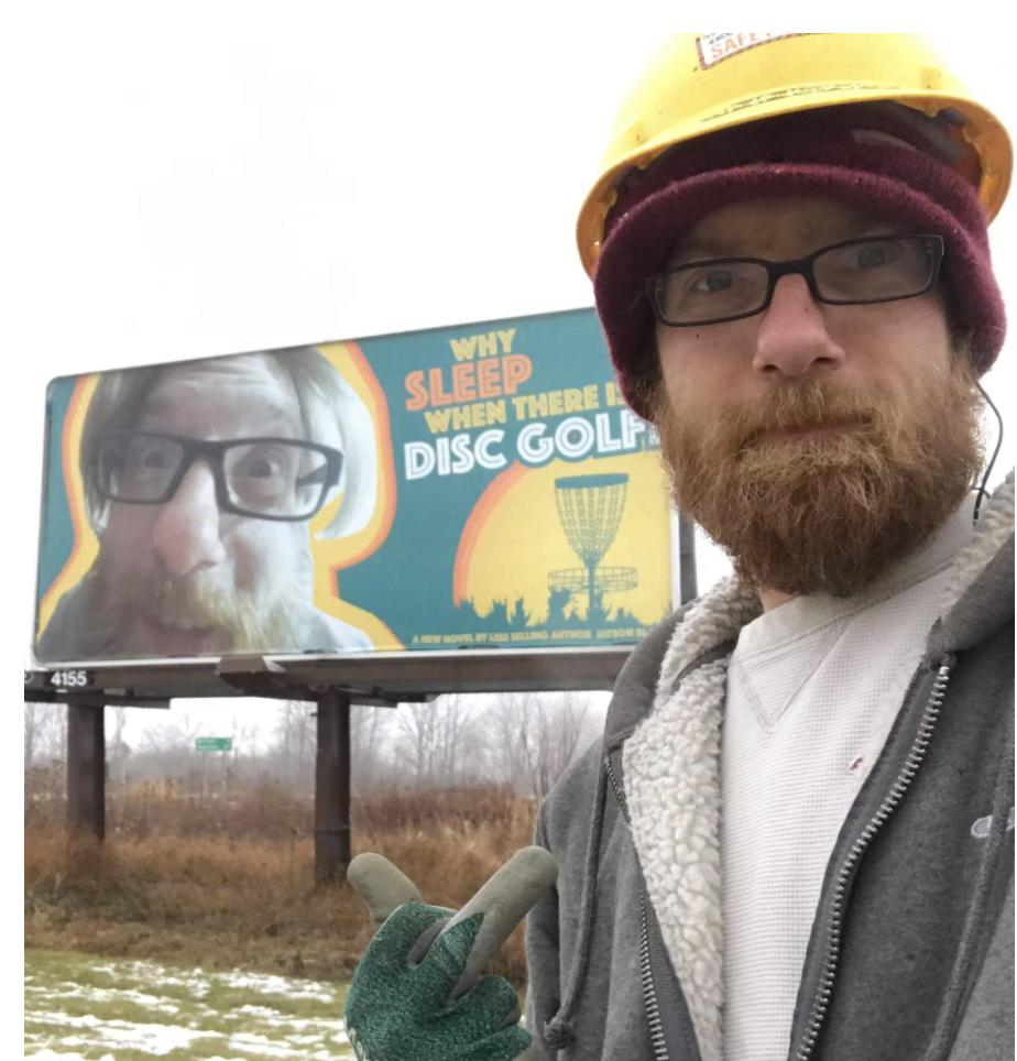 An annoyed man gives the camera the finger as he stands in front of a giant billboard with his face on it reading: Why sleep when there is disc golf