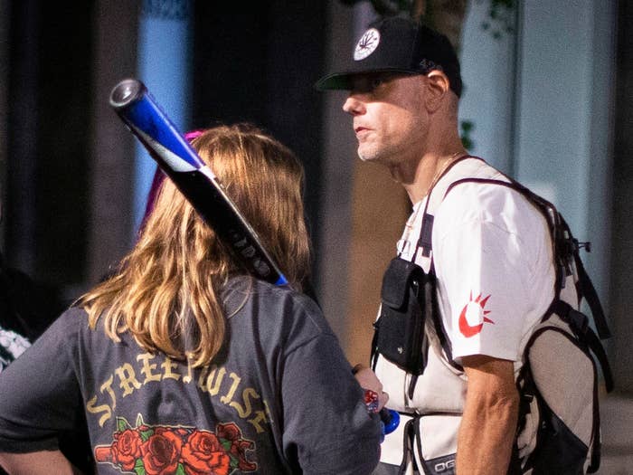 A tall man wearing a baseball cap stands next to a person with long hair holding a baseball bat whose back is to the camera.