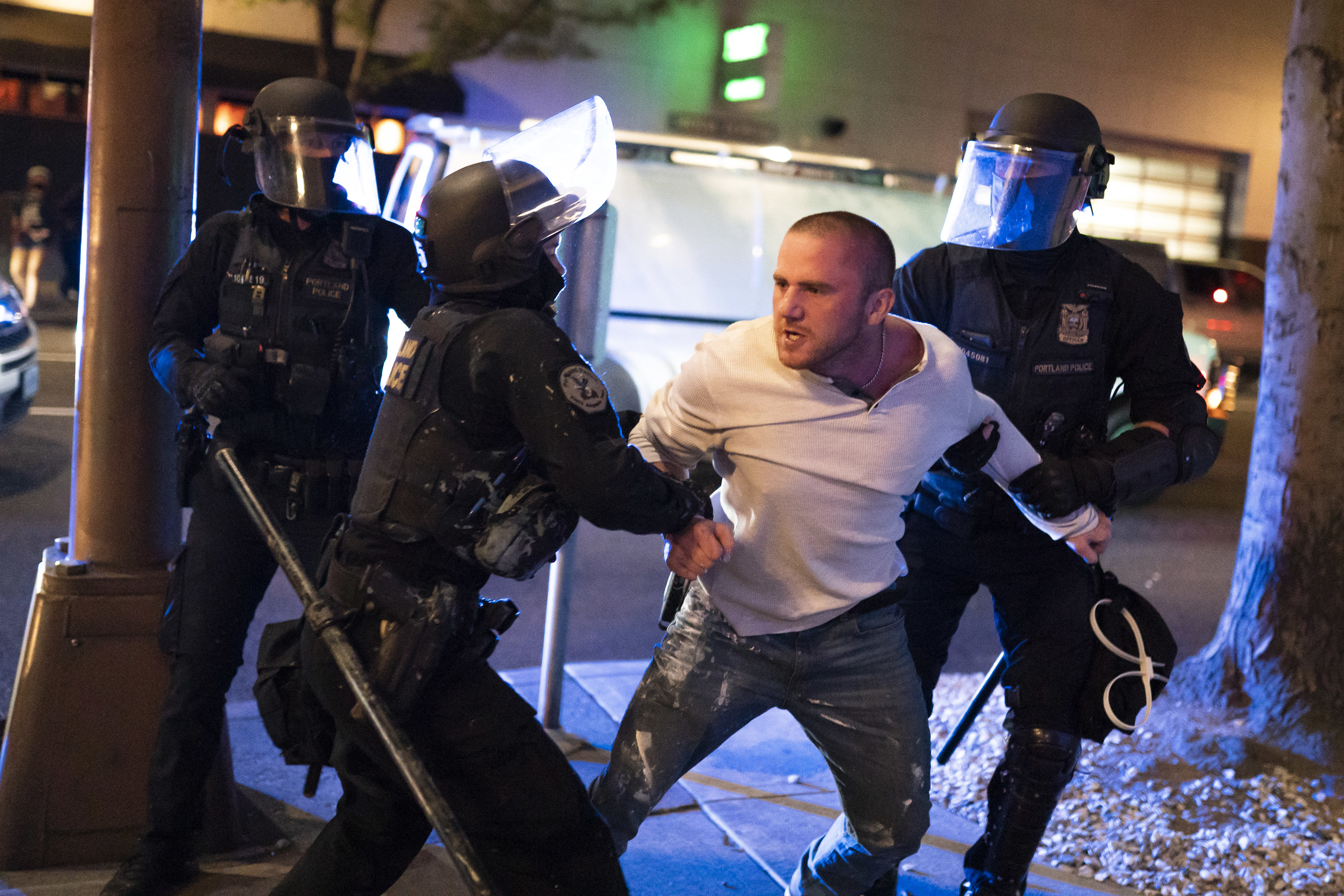 A man looks shocked as he is restrained by three officers wearing riot gear.