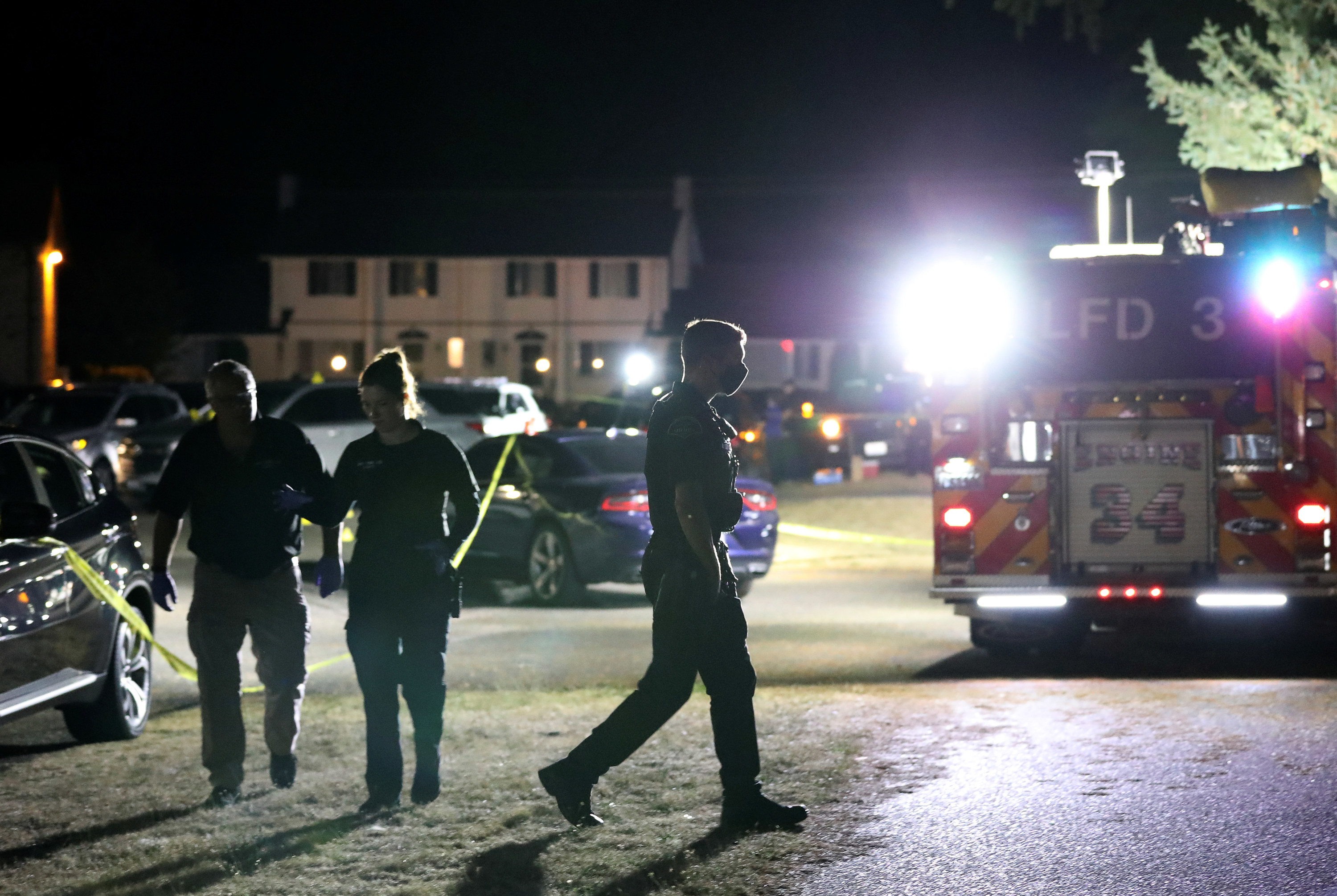 Three people walk among cars as a fire truck shines its lights at the camera.