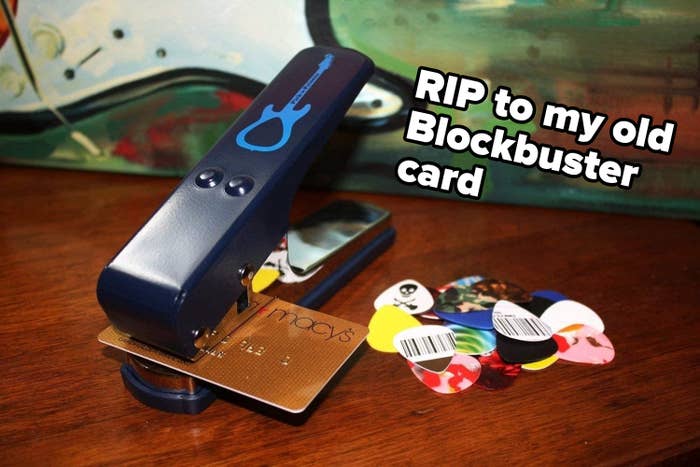 The guitar pick punch getting ready to make a pick out of a Macy&#x27;s card sitting next to a pile of picks made from various cards and plastic with the caption: &quot;RIP to my old Blockbuster card&quot;