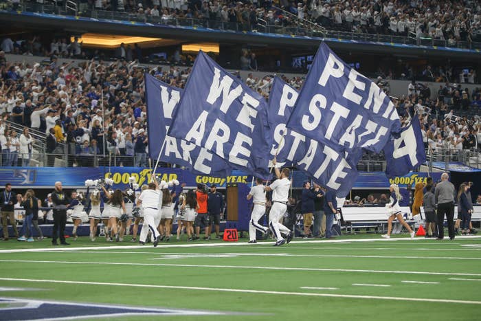 Four men run on grass in a packed stadium as they hold giant flags that read &quot;We Are Penn State&quot;