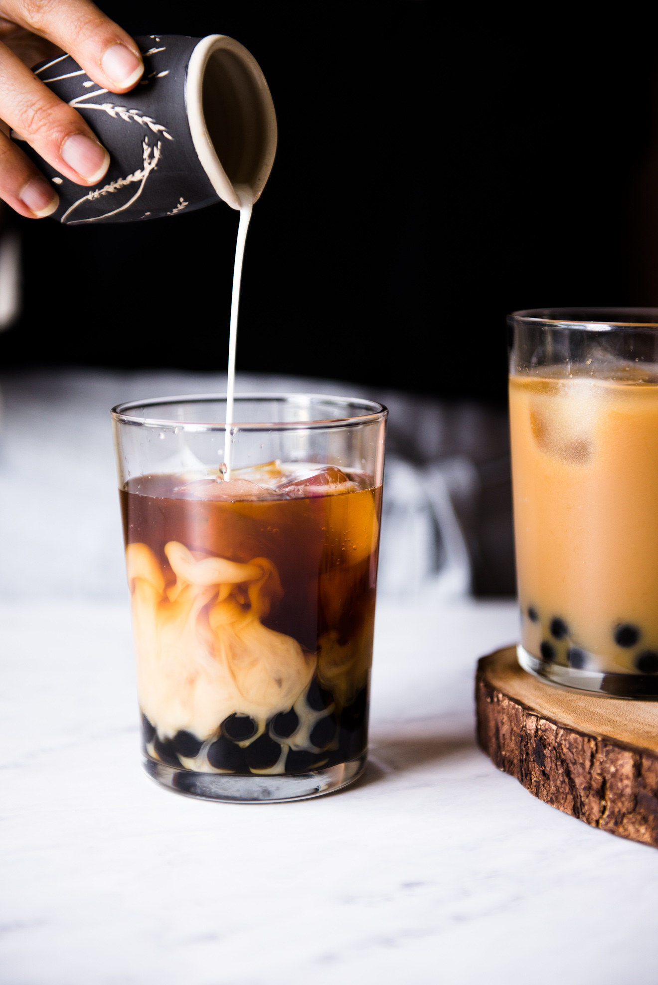 A hand pours a thin stream of milk into a clear glass in which it mixes with black tea and boba pearls for a swirling effect