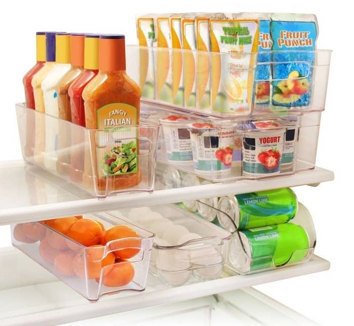 Six clear bins in a refrigerator filled with salad dressings, fruit, soda, yogurt, juice, and more