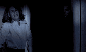 Laurie is resting by the dark doorway and Michael Myers&#x27; mask slowly comes into view