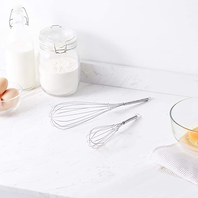 Two whisks on a white table along with a bottle of milk, a jar of flour and some eggs in a bowl