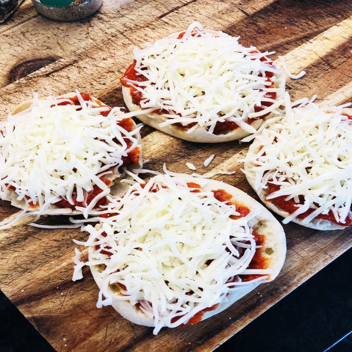 Four English muffins on a cutting board topped with tomato sauce and shredded cheese.