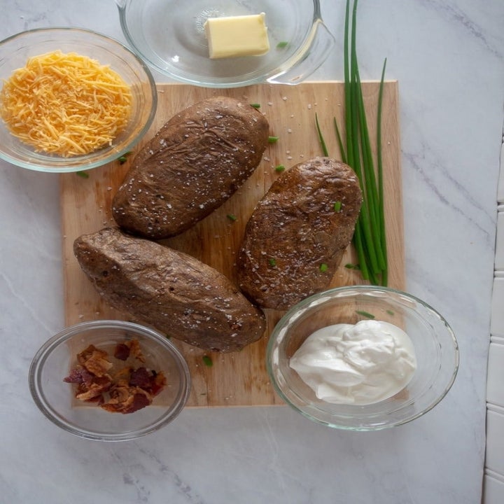 Ingredients for twice baked potatoes on a cutting board including potatoes, shredded cheddar, sour cream, bacon bits, butter, and scallions.