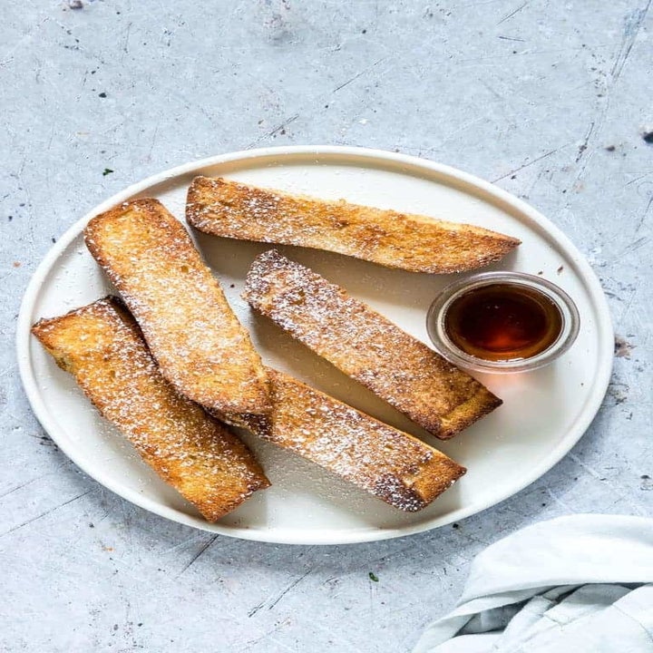 Five French toast sticks topped with powdered sugar on a plate with a side of maple syrup for dipping.