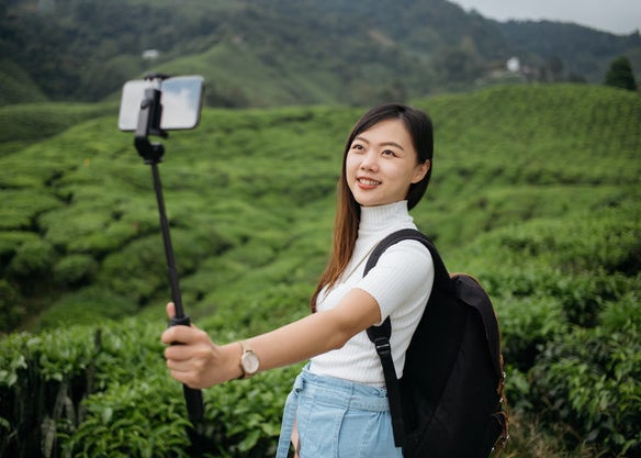 A woman poses for a photo with her selfie stick in front of lush, green fields