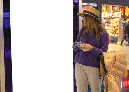 A woman in a hat, chunky sweater, and sweats looks to the side as a suitcase topped with a puffy coat sits next to her