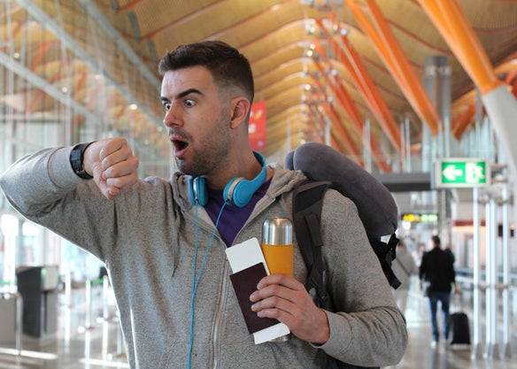 A man dramatically looks at his watch with a shocked face and a drink and passport in hand