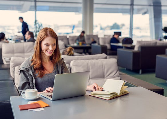 A woman smiles at a table in an airport surrounded by her laptop, open notebook, and a coffe drink