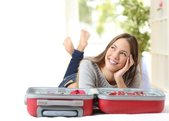 A woman lays on a bed in front of an open suitcase, head in hands and smiling off behind the camera