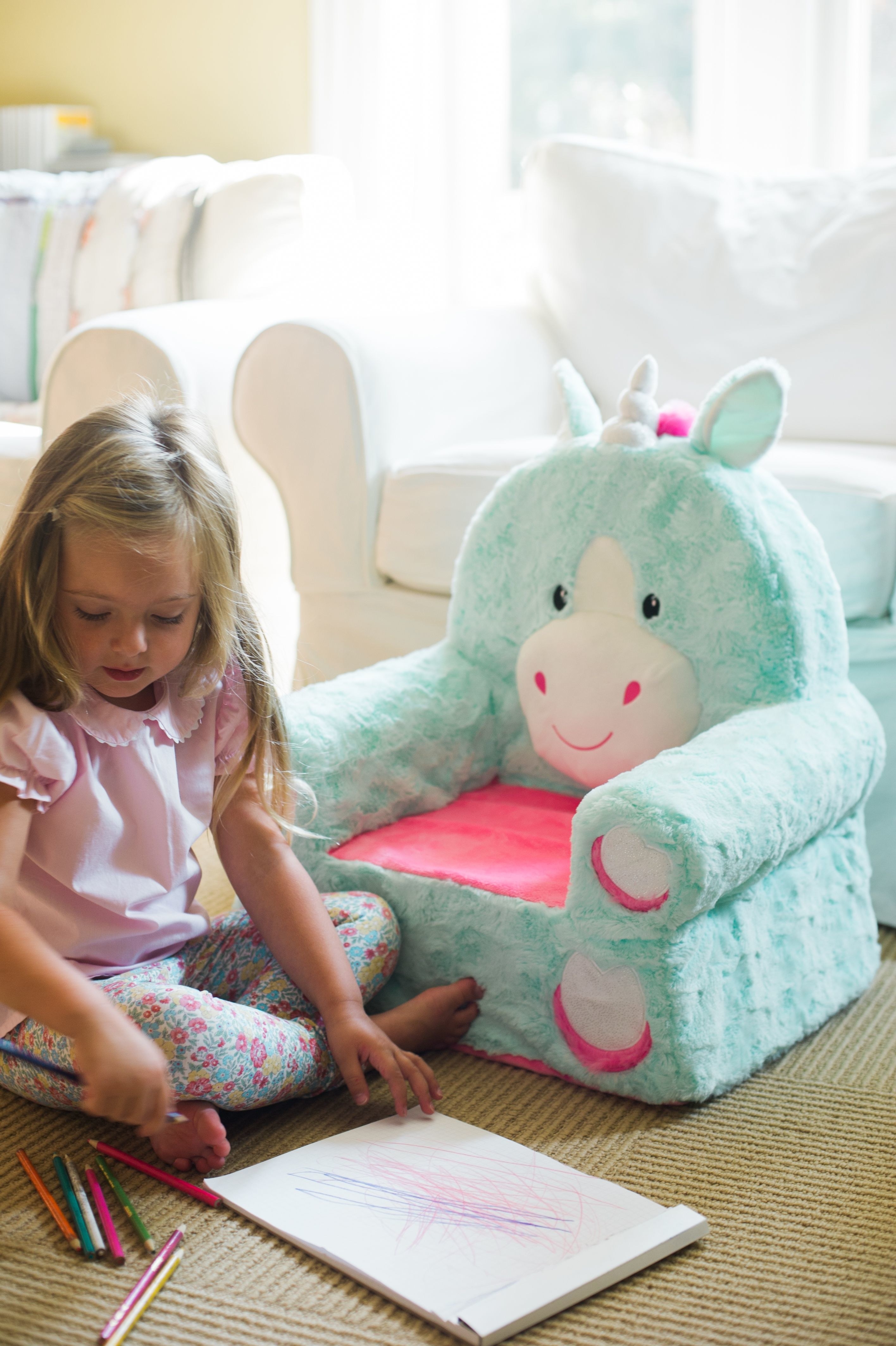 A turquoise and pink plush unicorn chair next to a child