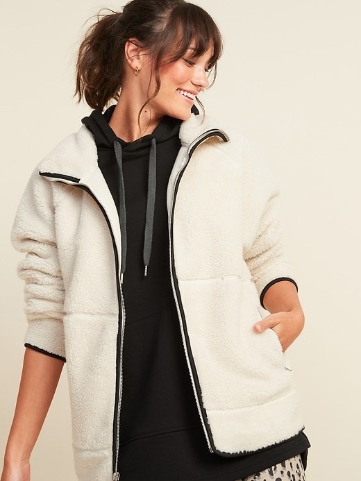 Model wearing the white fuzzy jacket with black trip, zipper up the front, and large pockets on the sides.