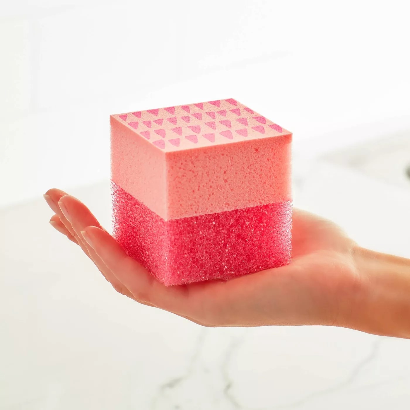 A model holding a pink, cube-shaped sponge with a soft side and a scrubby side