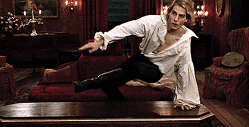 Lestat hops over a coffin and pats the space next to him