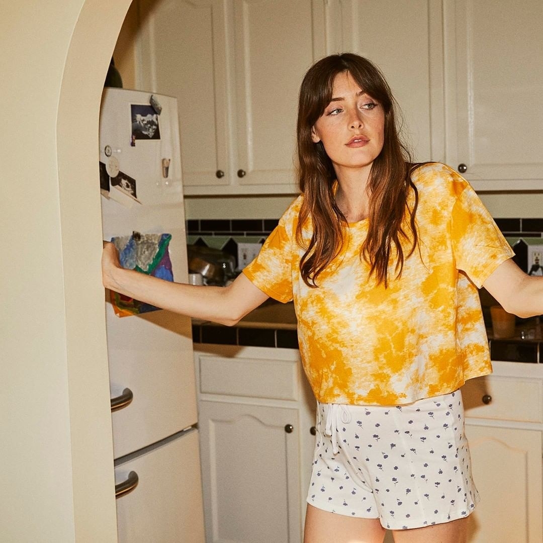 A model wears a yellow and white tie-dye, flowing, cropped t-shirt as she looks out from her kitchen