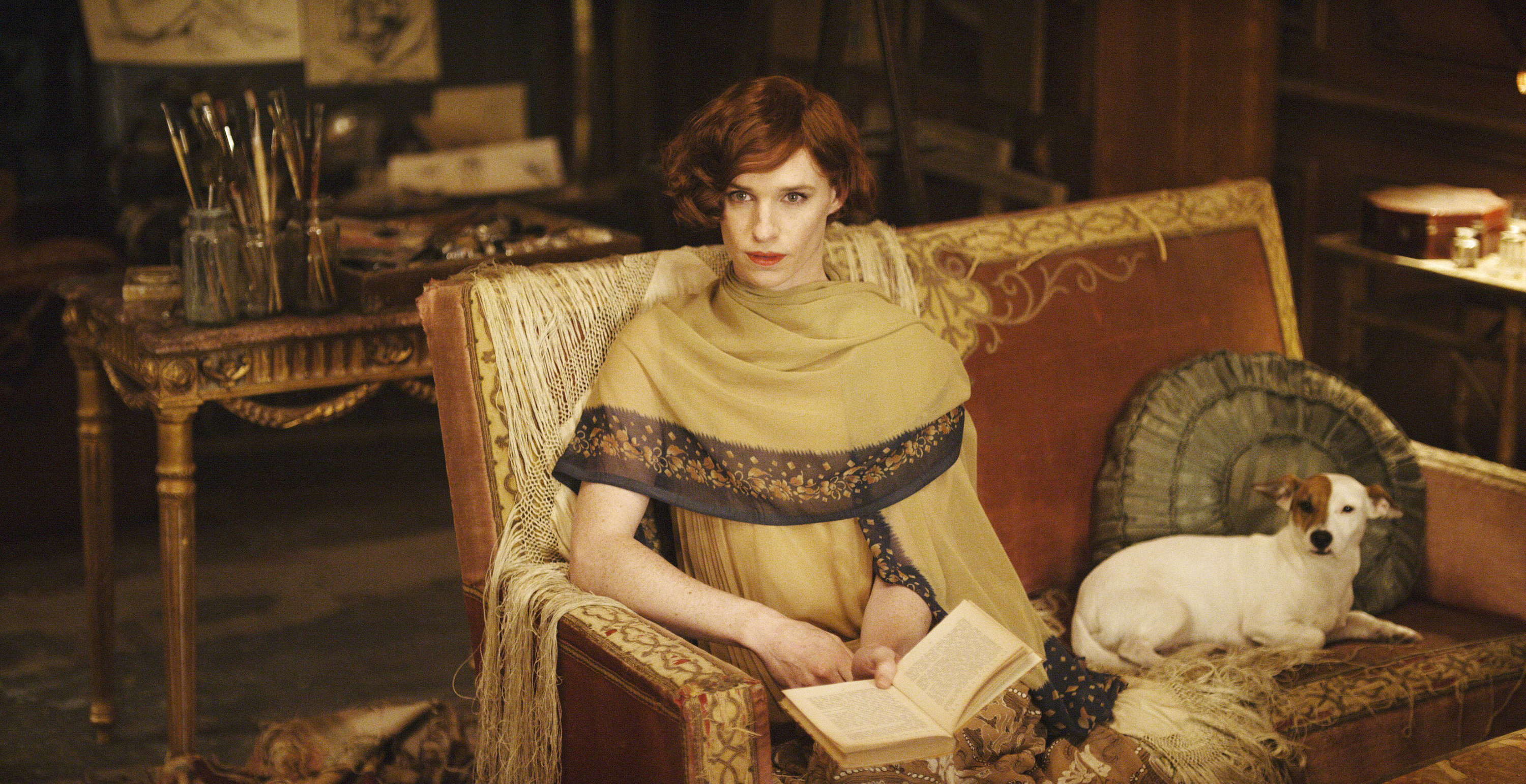 Eddie Redmayne as Lili Elbe, reading in a dress on her couch next to her dog. 