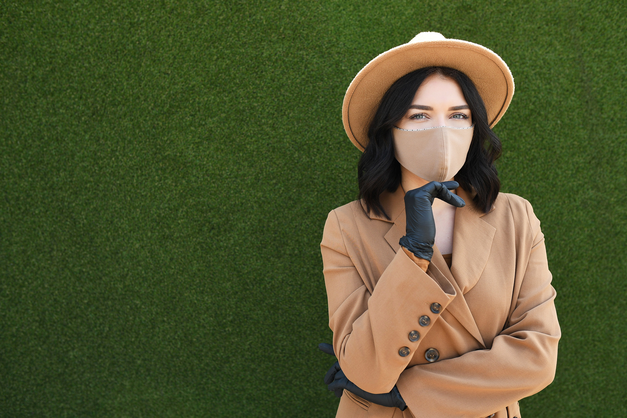 Une personne qui porte un masque assorti à son manteau et à son chapeau. Elle porte des gants en cuir et ses bras sont repliés. La photo est prise sur un fond herbeux.