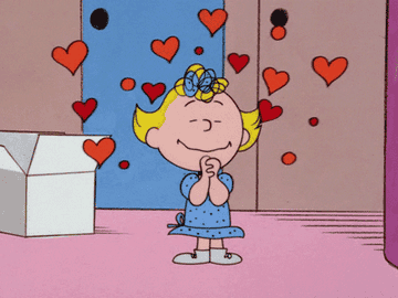Sally Brown holding her hands together and looking happy; there are floating love hearts animated around her