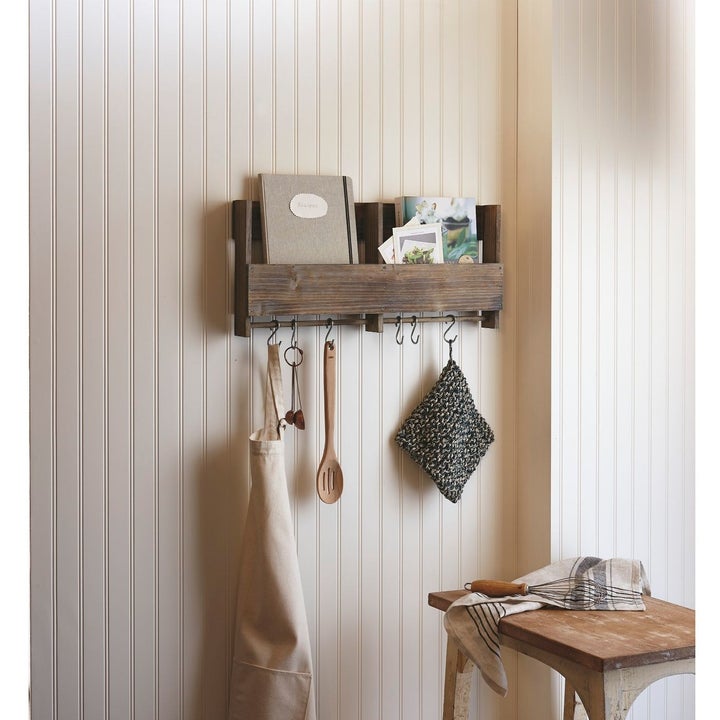 The shelf mounted on a wall, holding several books inside its shelved area, with wooden spoons, pot holders, and an apron dangling from the hooks